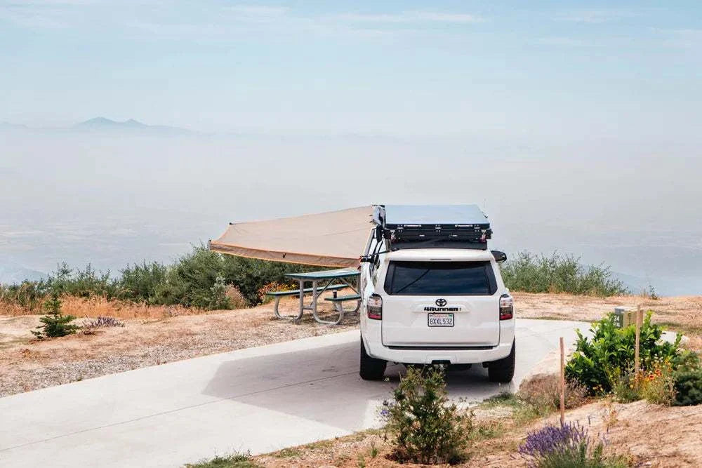 Outdoor scene with a Tuff Stuff® Overland 180 Degree XL awning attached to a white SUV, providing shade at a scenic overlook.