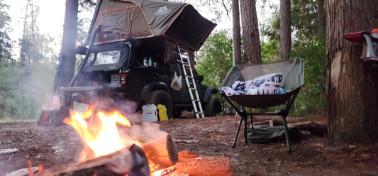 Rugged TUFF STUFF® Overland Stealth Black Ops Series aluminum shell RTT set up on a vehicle in a forest campsite with tent open, ladder attached, and campfire burning.