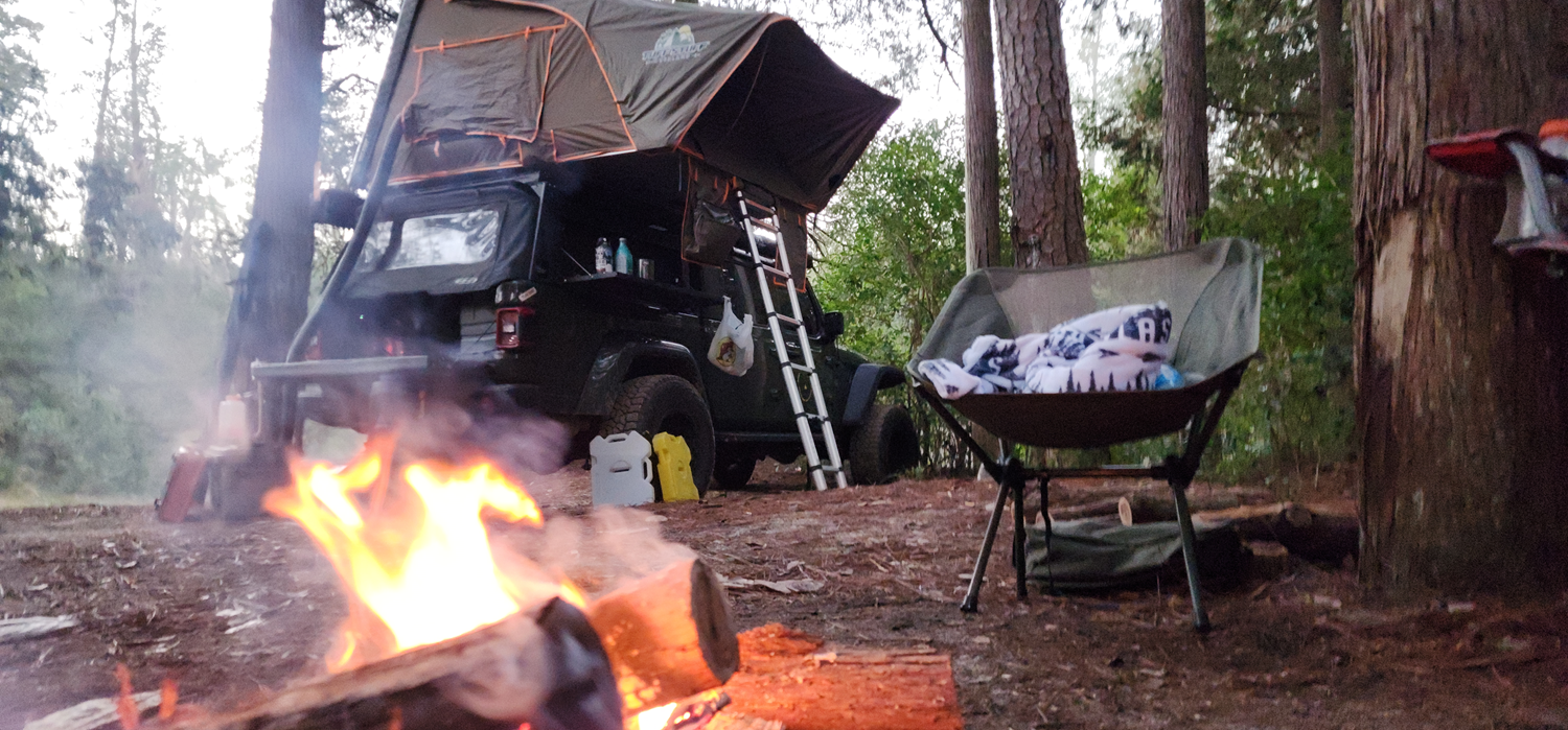 Rugged TUFF STUFF® Overland Stealth Black Ops Series aluminum shell RTT set up on a vehicle in a forest campsite with tent open, ladder attached, and campfire burning.
